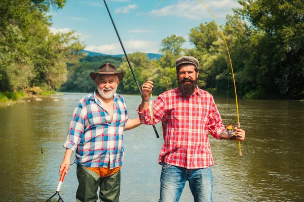 Aktivita a koníček. Nastavte tyč s navijákem. Dva mužští přátelé oblečení v košilích rybaří spolu se sítí a prutem během ranního světla na jezeře. Portrét veselého staršího muže rybaření. — Stock fotografie