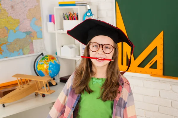 Retrato de colegial engraçado com caneta na boca em chapéu de formatura em sala de aula. Criança estudando. — Fotografia de Stock