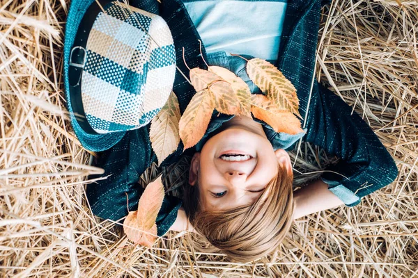 Leende pojke leker med löv och tittar på kameran ligger på höet. Söta lilla barn på gården byn. Barndom på landsbygden. — Stockfoto