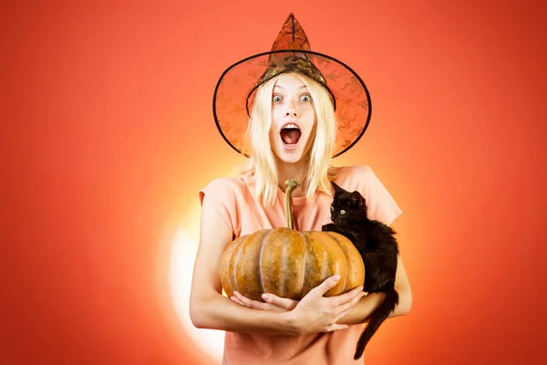 Dziewczyna Halloween. Kobieta w kapeluszu czarownicy na pomarańczowym tle. Portret pięknej blondynki trzymającej dynię i czarnego kota na pomarańczowym tle. — Zdjęcie stockowe