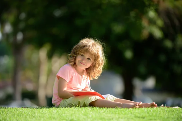 Förskolan ler elev pojke skriva till anteckningsbok i parken på gräs. Hemma lär sig barn. Grundläggande läxskolekoncept. — Stockfoto