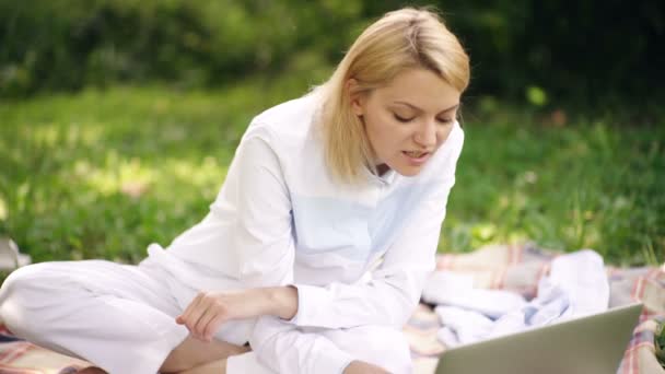 Una chica en un césped con un portátil. Estudiante chica está haciendo la tarea a través de la netbook mientras está sentado en el prado. — Vídeos de Stock