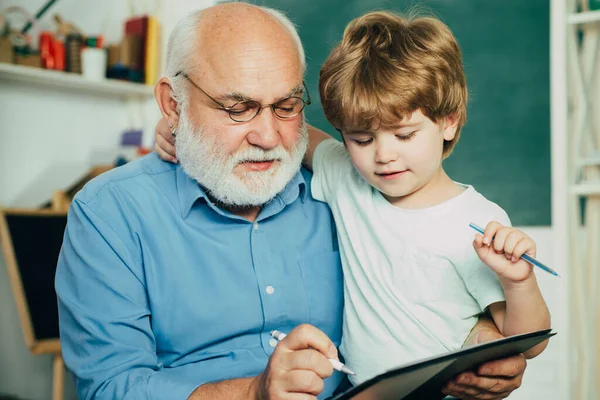 Großvater und Enkel lernen zusammen. Freundlicher Junge mit alter, reifer Lehrerin im Klassenzimmer neben der Schultafel. Lehrer ist kompetenter Anführer. — Stockfoto