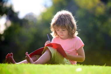Komik çocuk ödevini yapıyor ve parkta çim üzerinde kitap okuyor. Çocuklar için uzaktan eğitim. Anaokulu öğrencisi.