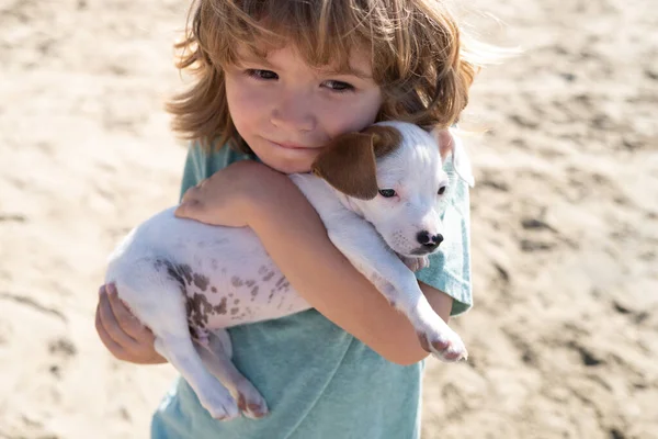 Ευτυχισμένο αγόρι αγκαλιάζει το σκύλο του στην άμμο. Το παιδί αγκαλιάζει με αγάπη το σκυλάκι του. — Φωτογραφία Αρχείου