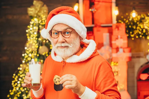 Santa Claus má rád sušenky a mléko vynechané na Štědrý večer. Veselý Santa Claus drží sklo s mlékem a sušenkou s krbem a vánoční strom v pozadí. — Stock fotografie