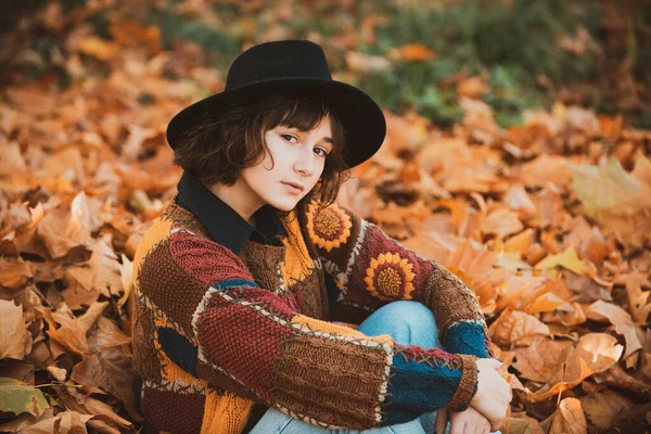 Mulher jovem hipster em uma camisola de malha na moda enquanto se senta na folhagem de outono na floresta. Modelo de outono. Maquiagem brilhante. Modelo da menina na temporada de outono. — Fotografia de Stock
