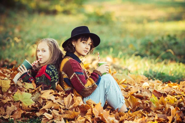 Tempo quente e ensolarado. Beleza mulher saudável e natural. Mulher sorridente alegre com irmã segurando xícara com chá quente ou suco. Meninas de outono brincando no parque. Retrato de irmãs no parque em cores de outono. — Fotografia de Stock