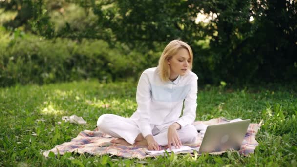 女生在室外。女孩坐在公园里的毛毯上，带着笔记本电脑工作。在户外学习的年轻妇女. — 图库视频影像