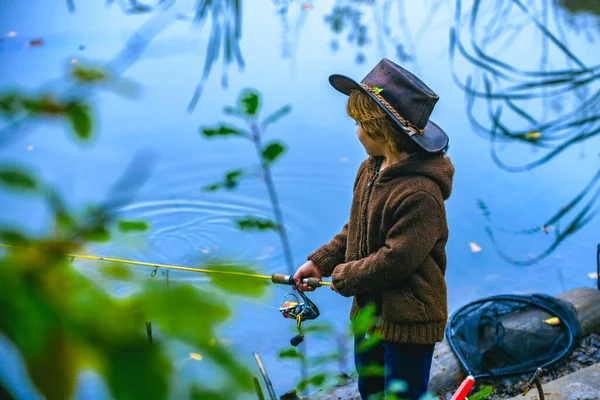 Ragazzino che pesca in tuta da un molo sul lago o stagno. Bambino con una canna da pesca in piedi vicino all'acqua. Giovane pescatore su un viaggio di pesca, vista posteriore. — Foto Stock