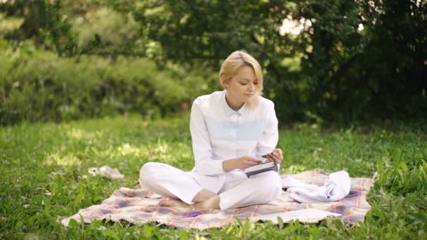La chica toma notas en un cuaderno sentado en una manta en el parque. Estudiante estudiando en un parque. — Vídeo de stock