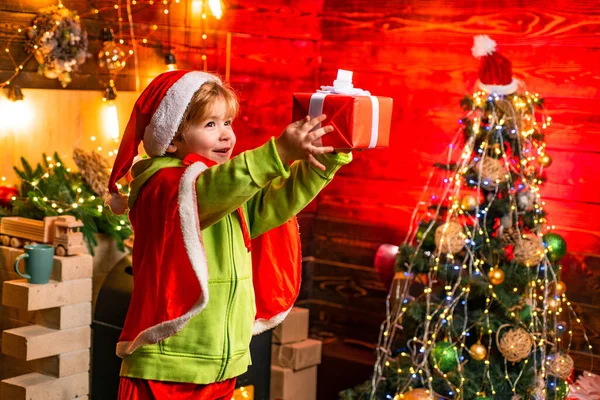 Dziecko trzyma pudełko na choinkę i tło kominka w Wigilię Bożego Narodzenia. Atrybuty świąteczne. Świąteczne zakupy. Cute wesoły dziecko gra w pobliżu choinki. — Zdjęcie stockowe