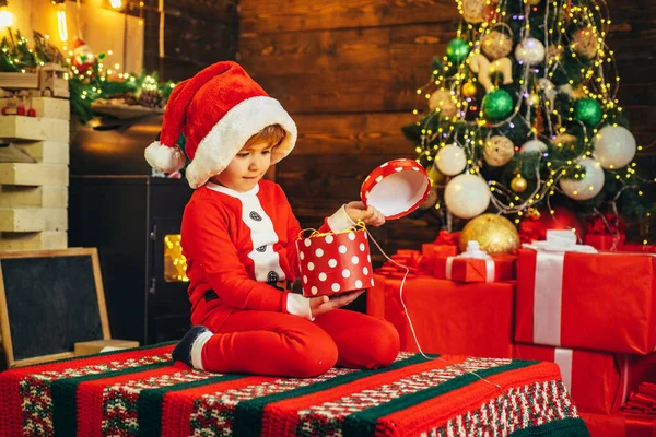 子供はサンタの服を着ていて、クリスマスプレゼントをクリスマスライトで開けています。クリスマスのコンセプト。サンタ・ハット. — ストック写真