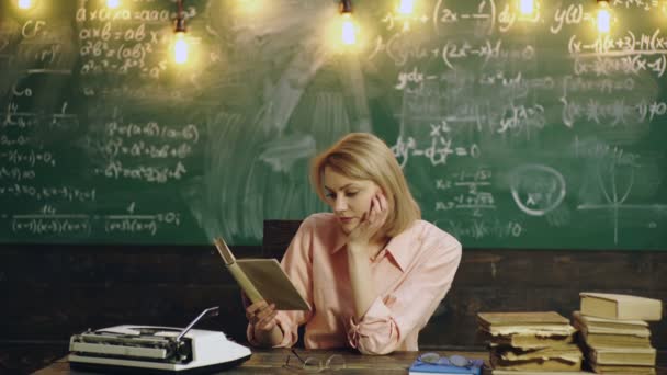 Enseignante assise dans la salle de classe tout en lisant et en écrivant sur le livre. Étudiant étudiant près du tableau noir devant sur le tableau noir. — Video