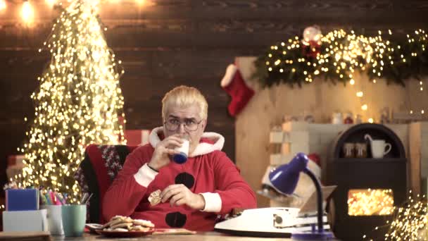 Der moderne Weihnachtsmann pflückt Plätzchen und ein Glas Milch und schaut fern, wenn er zu Hause die Fernbedienung hält. Heiße Weihnachten noel. — Stockvideo