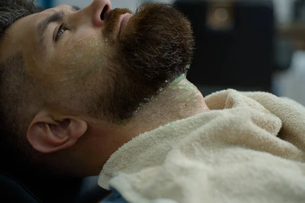 Bearded man getting haircut by hairdresser at barbershop. Razor sharp. Sandalwood shaving cream. Balm on dry body parts to moisturize skin. Old fashioned vintage barber shop pole.