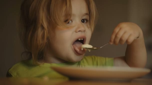 Kleine knappe blonde jongen eet een pap. Kind eet pap zelf met lepel en kijkt tekenfilms — Stockvideo