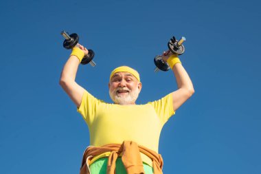 Old man holding his hands in front of him while lifting dumbbells. Senior man exercising on blue sky. Senior sportsman in sport center. clipart