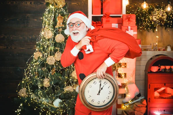 Weihnachtsmann. Porträt des überraschten und lustigen Weihnachtsmannes. Weihnachtsmann mit Wecker. Grußkarte Weihnachten. Foto vom Weihnachtsmann, der auf die Uhr zeigt, die fünf Minuten vor Mitternacht zeigt. — Stockfoto
