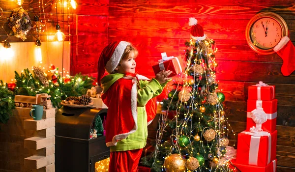 Noel ağacının yanındaki küçük Noel Baba birine Noel hediyesi veriyor. Küçük çocuk Noel Baba kıyafeti giyiyor.. — Stok fotoğraf