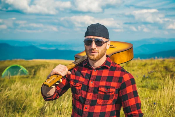 Caminante hombre con la guitarra mirando hermosa vista. Vacaciones de camping. Hombre hipster excursionista en las montañas. Retrato de hombre seguro de sí mismo. —  Fotos de Stock