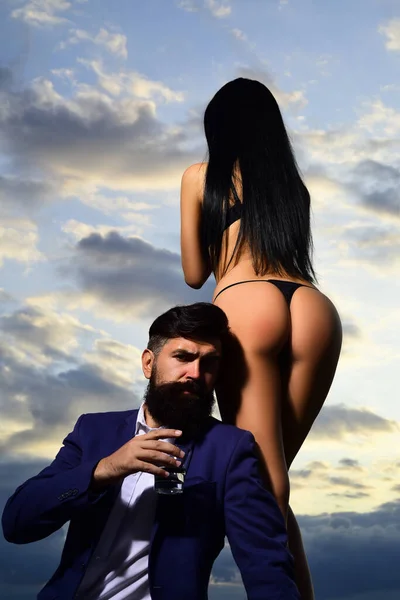 Bearded businessman in elegant suit with glass of whiskey. Rich man drink. Sencual couple in love. Womans butt in bikini. — Stock fotografie