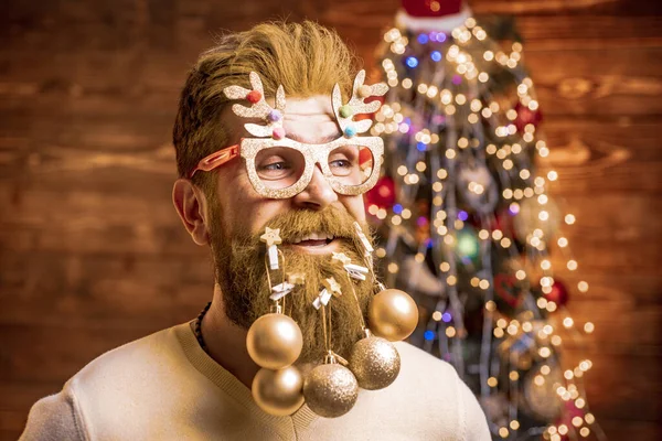 Бородатий чоловік розважається біля ялинки в приміщенні. Стилізація Санта-хіпстера з довгою бородою, що позує на ялинковому дерев'яному тлі. Зимові емоції . — стокове фото