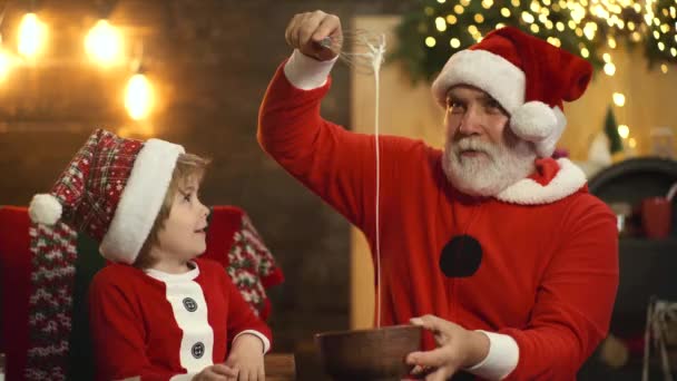 祖父と孫はサンタの帽子でクリスマスや新年を準備します。サンタと子供は面白い顔をし、ヴィンテージキッチンでクリスマスクッキーを焼く. — ストック動画