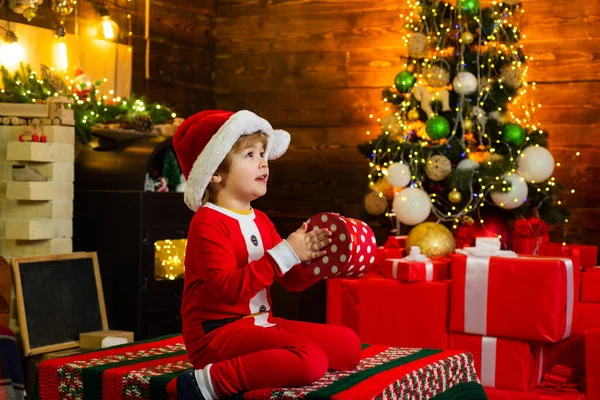 Conceito de Natal de Ano Novo. Criança feliz vestida com roupas de inverno pense no Papai Noel perto da árvore de Natal. Alegre bonito criança abrir um presente de Natal. — Fotografia de Stock