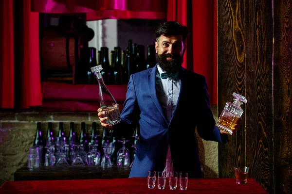 Luksusowy napój Barmana. Facet z brodą trzyma szkło z alkoholem w barze. Barman kelner w zabytkowej kamizelce z whisky lub szkocką na tacy. — Zdjęcie stockowe