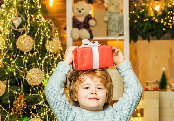 Criança engraçada segurando uma caixa de presente vermelha com ambas as mãos. Férias de Natal. Criança feliz vestida com roupas de inverno pense no Papai Noel perto da árvore de Natal. — Fotografia de Stock