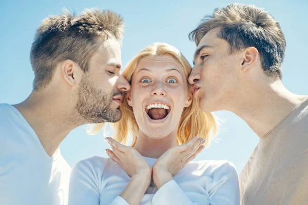 Blonďatá šťastná dívka dostává polibky od dvou pohledných chlapců. Okouzlující žena stojící mezi dvěma přáteli. Nejlepší přátelé. Skupina přátel na jasném pozadí oblohy. — Stock fotografie