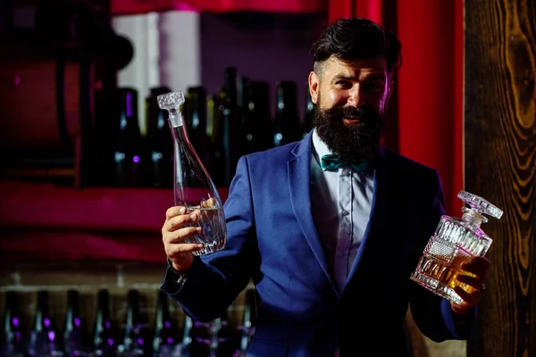 Handsome bearded barman with long beard and mustache with serious face made alcoholic cocktail in vintage suit on bar background.