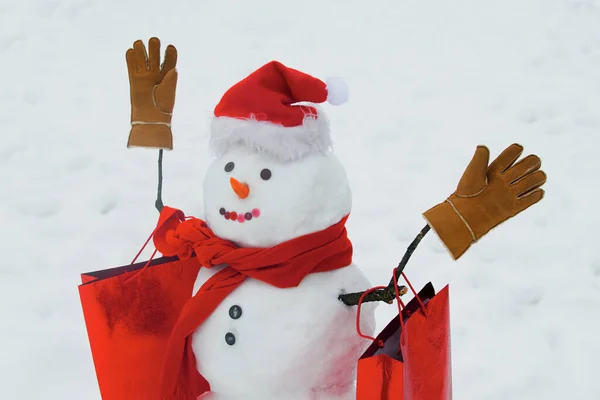 Feliz invierno. Regalos de entrega y emociones de regalo. Muñeco de nieve divertido con una zanahoria en lugar de una nariz y en un sombrero de punto caliente en un prado cubierto de nieve sobre un fondo de nieve borrosa. — Foto de Stock