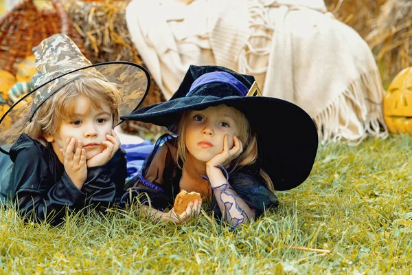 Gyerekek húga és testvére tökkel öltözve, csontváznak és boszorkánynak öltözve a Halloween partira. Boldog Halloween Gyermek lány és fiú ül széna vagy szalma a réten ősszel. — Stock Fotó