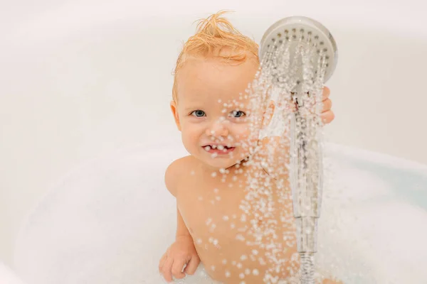 Csinos mosolygós kisfiú, aki szappanszappanos fürdőt vesz. Aranyos kisfiú vesz fürdő játszik hab és kacsa játékok egy fehér napos fürdőszobában. — Stock Fotó