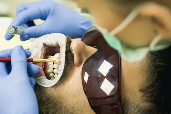 Słodki młody człowiek uśmiechnięty aparat fotograficzny. Dentystka sprawdzająca zęby pacjenta w nowoczesnej klinice stomatologicznej. Badanie młodego mężczyzny przez stomatologa na lekkim niewyraźnym tle. — Zdjęcie stockowe