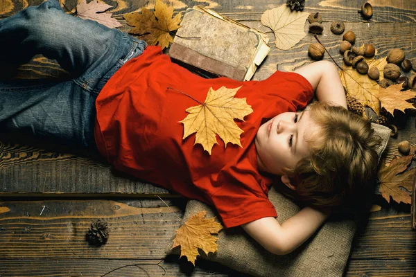 Маленький блондин, лежащий с листом на животе, лежит на деревянном полу в осенних листьях. Милый маленький мальчик готовится к осени. Ребенок веселится с Лиф Фолл. — стоковое фото