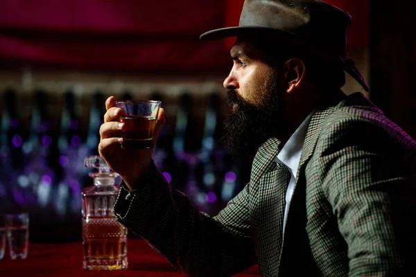 Handsome bearded barman with long beard and mustache with serious face made alcoholic cocktail in vintage suede leather on purple red bar background.
