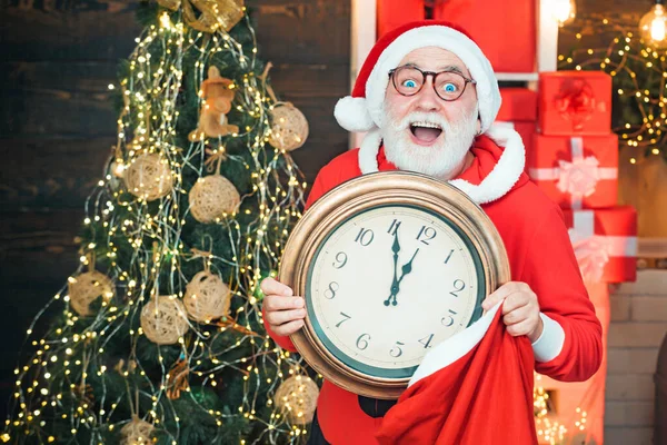 Weihnachtsmann hält Uhr mit Countdown zu Weihnachten oder Neujahr Weihnachtsmann in Holzhaus Interieur zeigt die Zeit auf einer Uhr. — Stockfoto