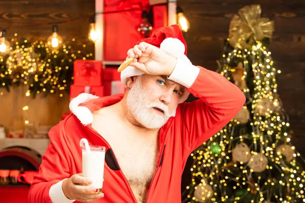 Alegre Papai Noel segurando vidro com leite e biscoito com lareira e árvore de Natal no fundo. Feliz Natal!. — Fotografia de Stock