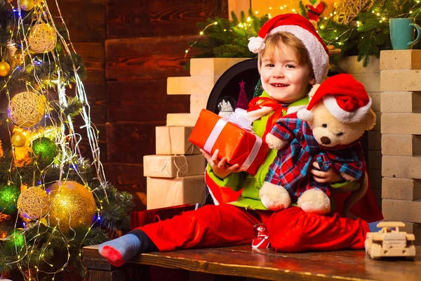 Neşeli, gülümseyen Noel Baba çocuk oyuncak ayısını ve Noel hediyesini şöminenin yanında tutuyor ve yeni yıl ağacını süslüyor. Küçük çocuk Noel ağacını süslüyor ve hediyeleri açıyor.. — Stok fotoğraf