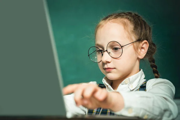 Alumno trabajando en el ordenador portátil sobre fondo pizarra. Niño cerca de pizarra en el aula de la escuela. El niño está aprendiendo en clase en el fondo de la pizarra. — Foto de Stock