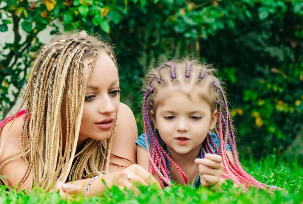 Lifestyle portrait mom and daughter in happy mood on grass. Happy mother hugging her daughter. Braiding hair. — Stock Photo, Image