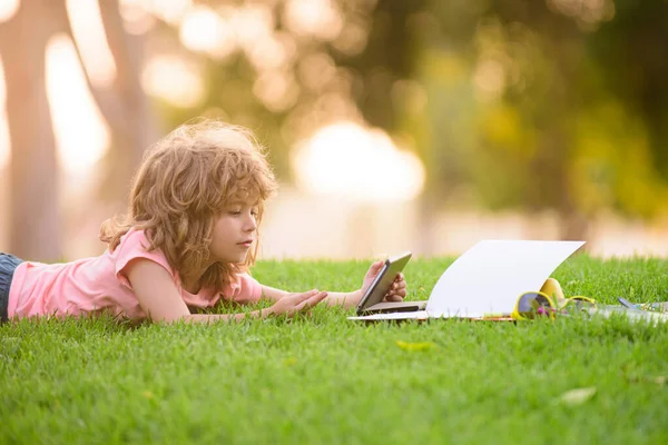 Dzieci studiują i uczą się z tabletkami na zewnątrz. Dzieciak ze szkoły zabawnej leży na trawie. Uczenie się na odległość. Uczeń chłopiec uczy się zdalnie na świeżym powietrzu w parku. Szkolnictwo na zewnątrz. — Zdjęcie stockowe