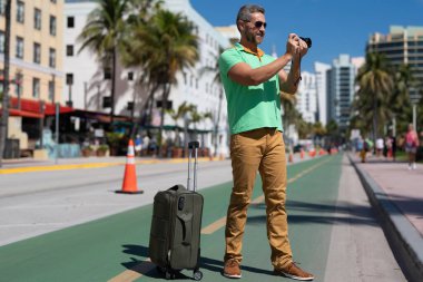 Miami sokaklarında fotoğraf çeken kameralı bir turist. Bir erkek turist, tropikal bir şehirde yaz tatilinin tadını çıkarır. Yaz şehrinde bavullu bir turist. Erkek turist şehirde yürüyor