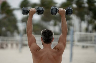 Spor adamının dambılları açık havada. Spor aktivitelerine katılan atletik bir adam. Formda adam geri döndü. Spor egzersizleri. Spor seansındaki üstsüz adam, geri döndü. Dambıllı açık hava eğitimi