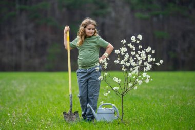 Çocuk dışarıda ağaç dikiyor. Bitkilerin bahar etkinliği. Çocuk kürek ve su bidonuyla çalışıyor. Çocuk meyve ağacı dikmiş. Bahçıvan çocuklar. Çocuk yetiştirme bitkisi ve ağaç bakımı. Bahar tarlasında küçük çiftçi