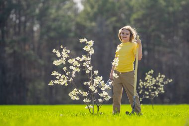 Çocuk bahçeye ağaç dikiyor. Çocuk kürekle toprak kazıyor ve ağaç dikiyor. Çocuk bahçıvan meyve ağaçlarıyla ilgileniyor. Çocuklarla ağaç dikmek. Yeşillik arayan bir çocuk. Çocuklar için bahçe.
