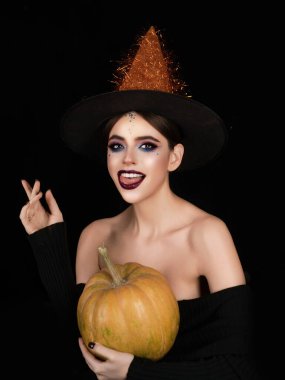 Kara stüdyoda balkabağıyla poz veren bir cadı. Cadılar Bayramı 'nı kutlayan cadı şapkalı genç kadın. Ürkütücü cadı kostümü portresindeki seksi model. Makyajlı ürkütücü, güzel cadı karakteri.